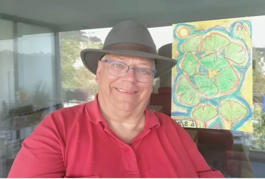 Beat Stricker malt immer das gleiche Motiv, und zwar Ginkgobäume mit einer imaginären Aura, einer Sonne und einem Vogel. Auf dem Bild ist Beat Stricker zu sehen, im Hintergrund eines seiner Bilder